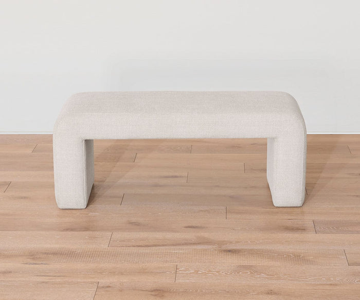 The Eilir Full Bench, featuring light beige upholstery and wide, block-style legs in a modern rectangular design, sits on a light wood floor against a plain white wall, offering extra seating with contemporary flair.