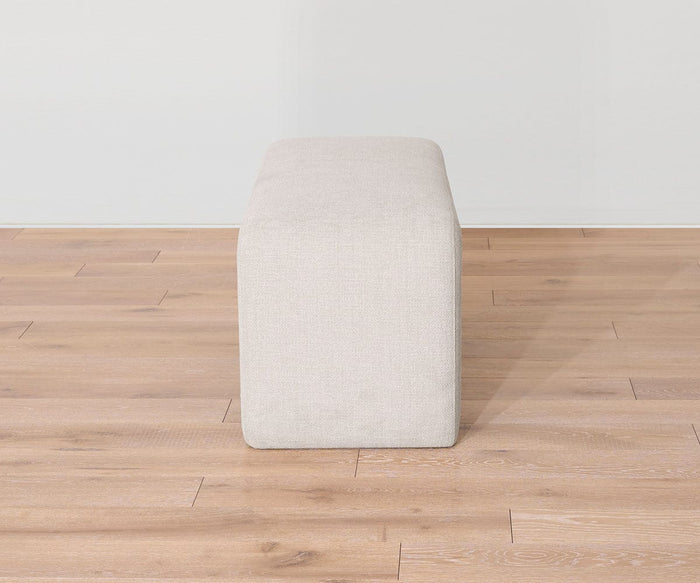 The Eilir Full Bench, a rectangular ottoman in light beige, sits on a wooden floor against a plain white wall and is viewed from the narrow end—perfect for adding extra seating and contemporary flair to any living space.