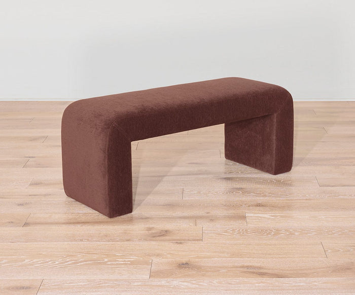 The Eilir Full Bench features a modern, smooth, and rounded design with a dark brown upholstered seat and legs, adding contemporary flair to any room. Placed on a light wood floor against a plain, light-colored wall, this bench offers both style and extra seating.