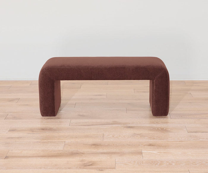 The Eilir Full Bench, featuring a modern design with a rectangular shape and rounded edges in a brown finish, sits on a light wood floor against a plain white wall, bringing contemporary flair and offering extra seating to the space.