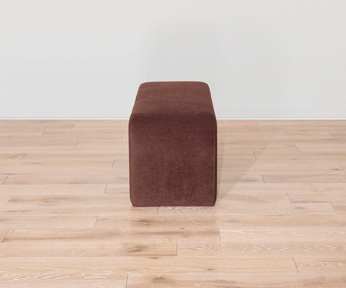 The Eilir Full Bench, in a rich brown finish, sits on a light wood floor against a plain white wall; viewed directly from the narrow end, it offers extra seating with contemporary flair.