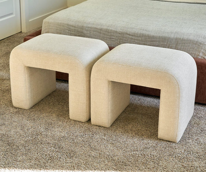 Two Eilir Half Benches, upholstered in beige with modern arch-shaped legs, sit side by side on a carpeted floor in front of a bed with a neutral bedspread, providing extra seating and adding contemporary flair to the room.