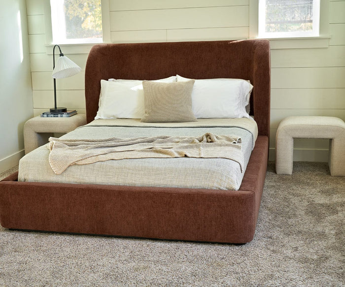 A modern bedroom features a brown upholstered bed with white and beige bedding, a small pillow, nightstands on each side topped with a lamp and books, and the Eilir Half Bench for extra seating—all set atop a beige carpet—for an added touch of contemporary flair.