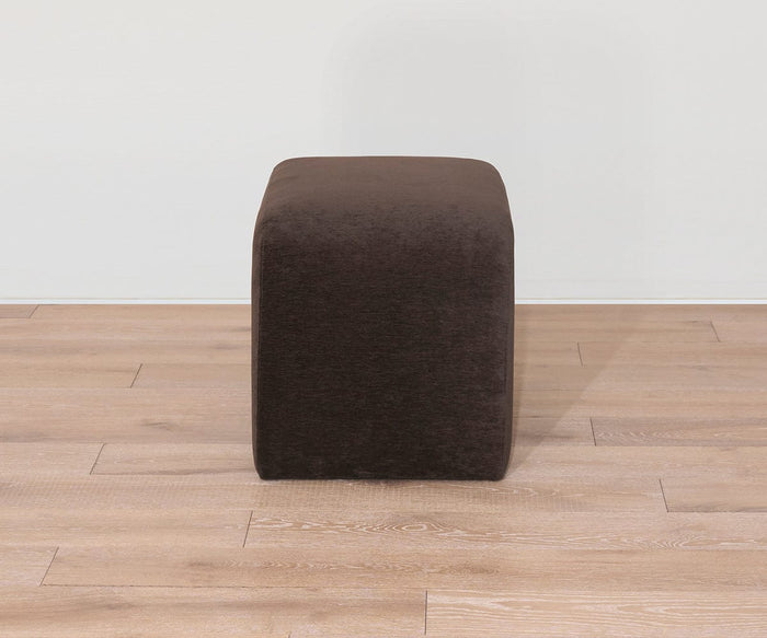 The Eilir Half Bench, a dark brown, cube-shaped ottoman, sits on a light wood floor against a plain white wall, offering extra seating with contemporary flair.