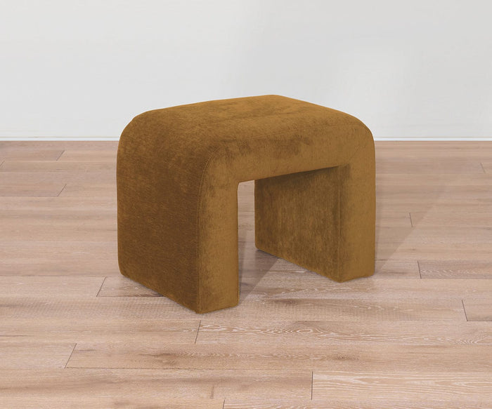 The Eilir Half Bench, a small, brown, velvet-covered piece with a modern arch-shaped design and contemporary flair, sits on a light wooden floor against a plain white wall.