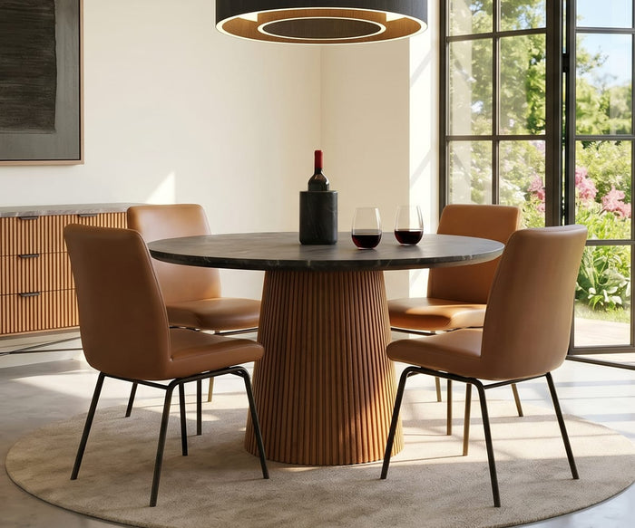 A modern dining room with a round table and four Elyse 1000 Dining Chairs in tan, each designed with subtle lumbar support for enhanced comfort. A wine bottle and two glasses sit on the table, while large windows fill the space with sunlight and offer views of lush greenery outside. A circular light fixture is suspended above the table.