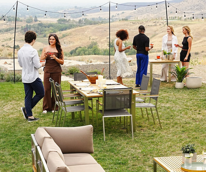 Six people chat and hold drinks around an outdoor dining table set on grass, with mountains in the background and string lights above. The table is surrounded by modern outdoor furniture, including a Farino Outdoor Dining Chair and a plush sofa, with food and drinks spread across it; some people stand while others sit comfortably.