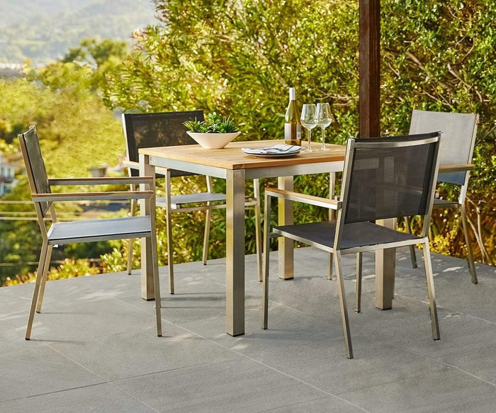 A modern outdoor dining set featuring a sleek square wooden table is complemented by four stylish Farino Outdoor Dining Chairs, perfect for al fresco gatherings. Two wine glasses, a wine bottle, and a bowl rest invitingly on the tabletop. Lush greenery and rolling hills provide a stunning backdrop, enhancing the appeal of this Modern Outdoor Furniture arrangement.