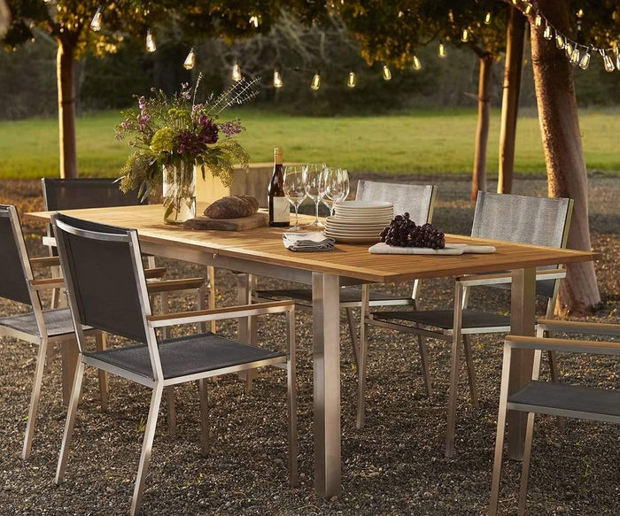 A wooden outdoor dining table set with plates, wine glasses, a wine bottle, bread, grapes, and flowers is surrounded by modern outdoor furniture, including sleek Farino Outdoor Dining Chairs under string lights and trees at sunset.