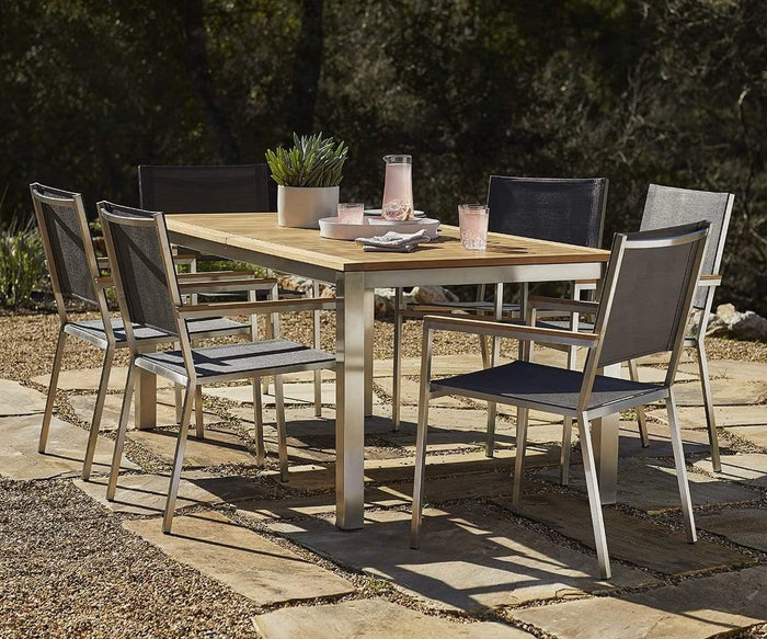 A modern outdoor dining table with six Farino Outdoor Dining Chairs in black and silver sits on a stone patio. The table, part of a stylish modern outdoor furniture set, features a potted plant, a glass pitcher of pink drink, and four glasses. Trees and sunlight create an inviting backdrop.