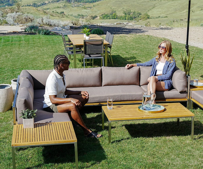 Two people sit on an outdoor sectional sofa in a sunny backyard, talking. Between them is the Farino Outdoor End Table, holding drinks and a plant; its solid acacia tabletop adds to the space’s modern design. A dining table with chairs sits in the background.