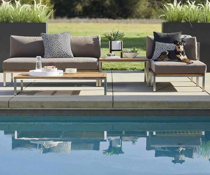 Modern outdoor lounge chairs with gray cushions sit by a pool, accompanied by the Farino Outdoor End Table featuring a solid acacia tabletop. Decorative pillows, a tray with drinks and snacks, and a dog resting on one chair add a cozy touch. Green plants and grass in the background enhance the fresh modern design of the space.