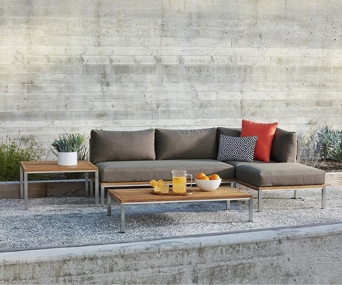 Modern outdoor patio set with gray cushions and the Farino Outdoor End Table, featuring a solid acacia tabletop, along with a planter, a jug of orange juice, glasses, and a bowl of oranges—all showcasing modern design against a concrete wall and gravel ground.