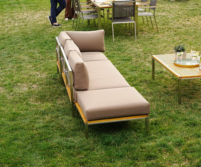 Outdoor patio featuring the Farino Outdoor Ottoman with beige UV-resistant cushions, a wooden table set with drinks and a potted plant, lush green lawn, and people gathered near another table with food in the background.