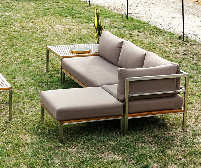 The Farino Outdoor Ottoman sits on a grassy lawn alongside a modern outdoor sectional sofa with taupe, UV-resistant fabric cushions, positioned next to a side table adorned with a plant and an ashtray.
