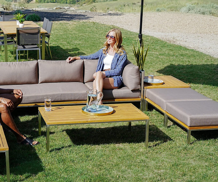 A woman wearing sunglasses and a blue striped blazer sits on the Farino Outdoor Ottoman with gray UV-resistant cushions, next to a table with drinks, on a grassy lawn with stainless steel furniture in the background.