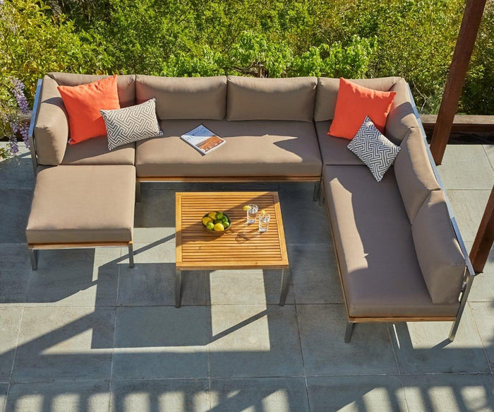 The Farino Outdoor Ottoman is featured on a tiled patio surrounded by greenery, paired with a beige sectional sofa upholstered in UV-resistant fabric. Four patterned and solid pillows add comfort, while a wooden coffee table holds lemons, two glasses, and a magazine for a relaxing outdoor setting.