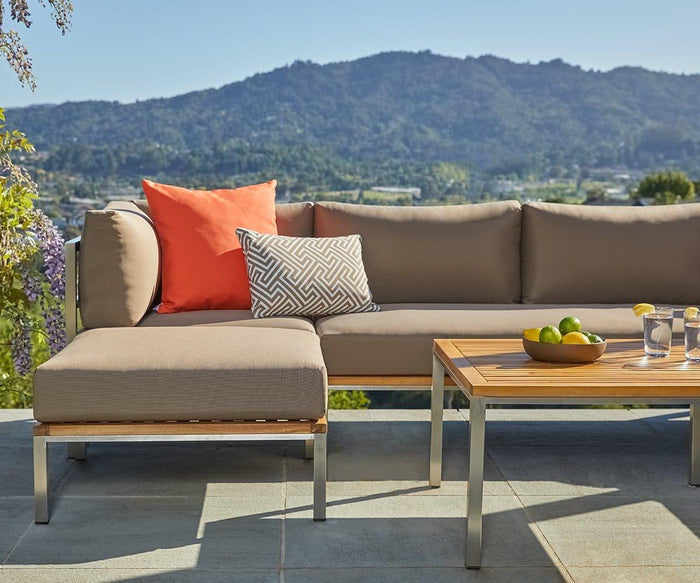 A modern outdoor patio features a beige sectional sofa with UV-resistant fabric, accented by an orange and a patterned pillow. A wooden coffee table with a bowl of limes and two glasses of water sits nearby, complemented by the Farino Outdoor Ottoman—all set against a scenic mountain backdrop.