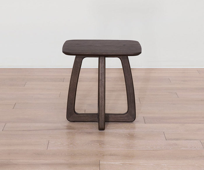 The Faroe Square End Table, featuring a small, square dark smoked oak veneer top and distinctive interlocking legs, is displayed on a light wood floor against a plain white wall.