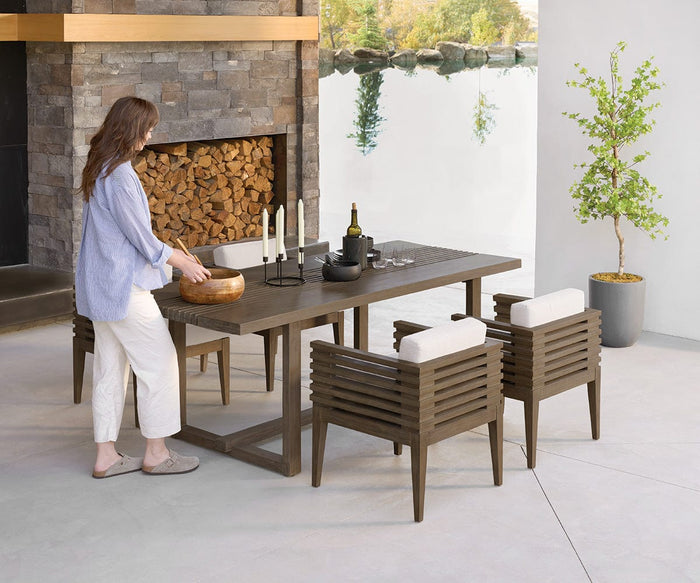 A woman arranges a wooden bowl on a modern outdoor dining table set with candles and dishes, surrounded by elegant Farren Outdoor Dining Chairs, near a stone fireplace and potted plant, overlooking a serene water view. Rewritten Sentence: A woman sets a wooden bowl on a contemporary outdoor dining table adorned with candles and dishes, while stylish Farren Outdoor Dining Chairs encircle the setting beside a stone fireplace and potted plant, all overlooking tranquil waters.