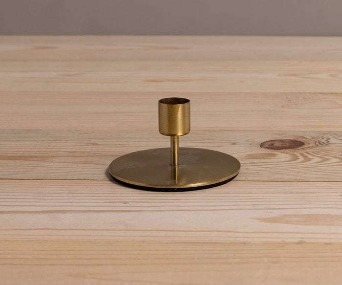 The Fina Candlestick, a minimalist handcrafted piece in gold with a round base, is placed on a light wooden surface against a plain, neutral background.