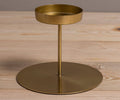 The Fina Pillar Candle Holder in gold features a round base and a raised circular tray supported by a vertical rod, displayed on a light wooden surface.