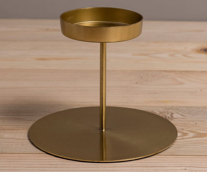 The Fina Pillar Candle Holder in gold features a round base and a raised circular tray supported by a vertical rod, displayed on a light wooden surface.