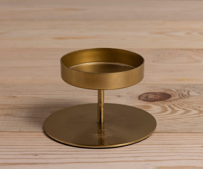 The Fina Pillar Candle Holder is a small, round, gold metal piece with a flat base and short stem, handcrafted and displayed on a light wooden surface.