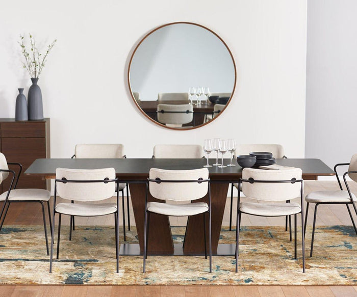 A modern dining room featuring a dark wooden table surrounded by eight beige Finn Dining Chairs, with a round mirror on the wall, vases filled with branches, stacked bowls, and wine glasses arranged on the table—all atop a patterned rug.