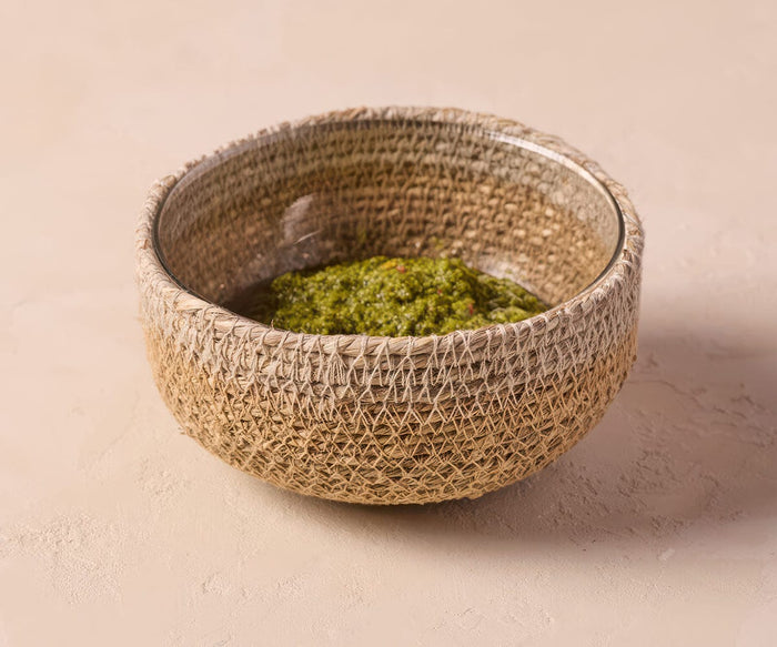 The Fjora Woven Bowl, handcrafted in India with a grey seagrass design, is filled with vibrant green pesto sauce and sits elegantly on a light beige surface.