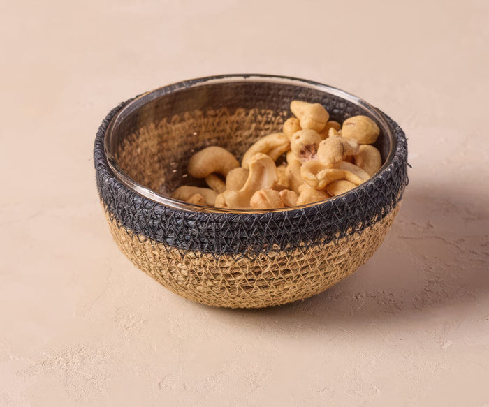 A navy Fjora Woven Bowl, handcrafted in India, is filled with cashew nuts and sits on a light beige textured surface.