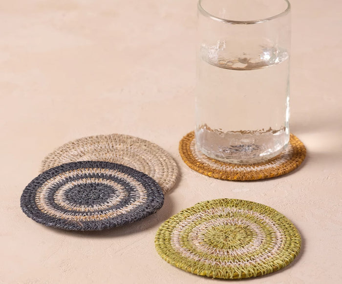 A clear glass of water sits on a round Fjora Woven Coaster in navy, handcrafted in India. Three additional Fjora Woven Coasters in green, blue, and beige are arranged nearby on a light, neutral surface.