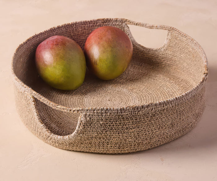Two ripe mangoes sit side by side in the Fjora Woven Tray, a round, grey tray with handles handcrafted in India, placed on a light beige surface.