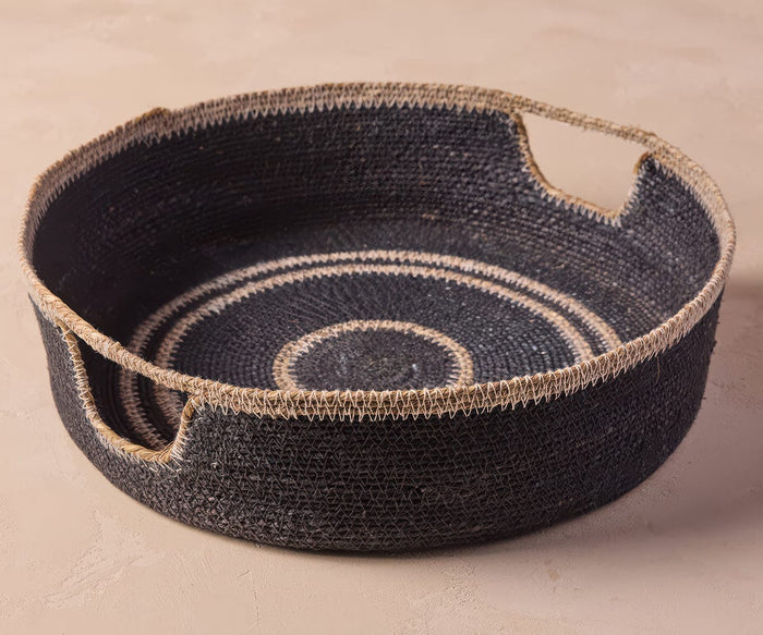 The Fjora Woven Tray, a round navy seagrass tray with beige trim and two handle cutouts, sits on a light beige surface. Handcrafted in India, the tray features intricate beige circular patterns inside.