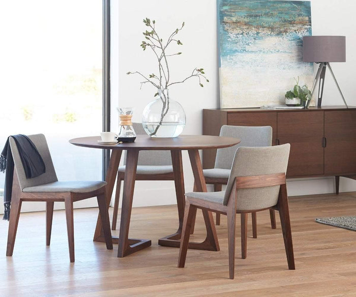 A round wooden table is paired with three Fuchsia Dining Chairs on a wooden floor. A glass vase with branches decorates the tabletop. Behind the ensemble, a sideboard stands beneath a lamp and an abstract painting on the wall.