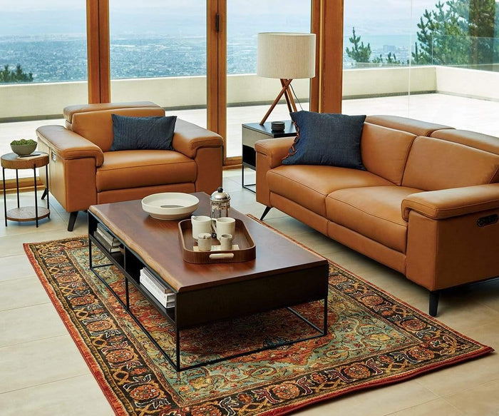 Modern living room with tan leather sofa and armchair, blue pillows, wooden coffee table with tea set, patterned rug, and large windows offering a scenic outdoor view. The Gaffel Table Lamp sits atop the side table, its tripod base adding a stylish touch that seamlessly complements the cozy home decor.