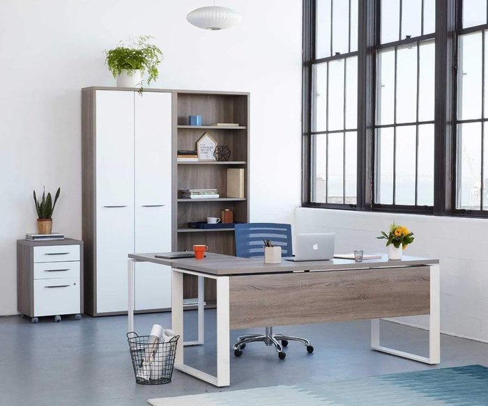 A modern office features a desk, chair, laptop, flowers, and neatly organized shelves. Large windows fill the space with natural light. Ample eco-friendly storage is provided by the Gammel High Bookcase Door Set with sleek white high-gloss doors. A potted plant and trash bin rest on the floor, complementing the minimalist and tidy décor.