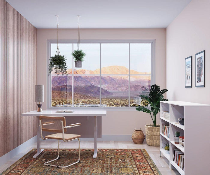 A modern home office features a desk, chair, bookshelf, houseplants, and a large window framing a mountain landscape. The space has light walls, wooden accents, and framed artwork on the wall, complemented by the Grazia Table Lamp—a mid-century style table lamp with an elegant hourglass-shaped base.