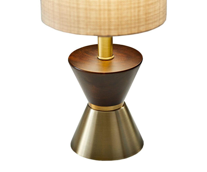 The Grazia Table Lamp features a modern mid-century design with a round beige lampshade, a wooden midsection, and a conical hourglass-shaped brass base, displayed against a plain white background.