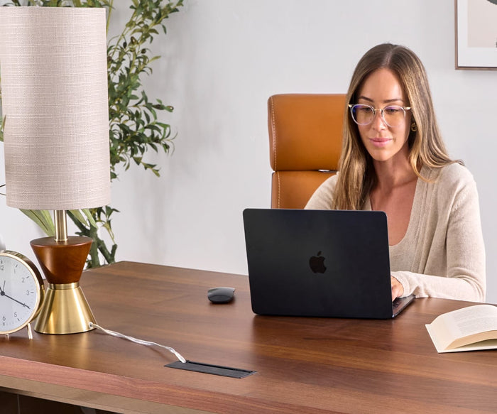 A woman wearing glasses works on a laptop at a wooden desk, illuminated by the Grazia Table Lamp with its hourglass-shaped base. Nearby, a clock, plant, and an open book complete the stylish workspace.