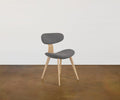 The Hana Dining Chair in Grey/Oak, featuring solid white oak legs and a gray performance fabric upholstered seat and backrest, sits on a brown floor against a plain white wall.