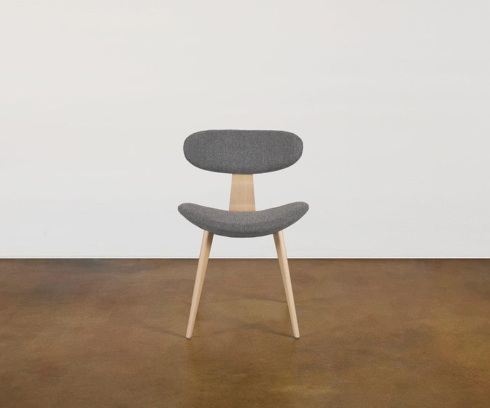 The Hana Dining Chair features a modern Scandinavian design with two solid white oak legs, a curved gray performance fabric seat, and a matching rounded gray backrest, set against a white wall and brown floor.