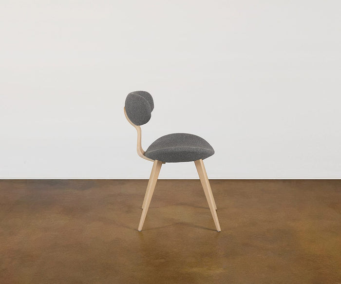 The Hana Dining Chair features curved solid white oak legs, a rounded gray seat upholstered in performance fabric, and a matching oval backrest, shown against a plain white wall and brown floor.
