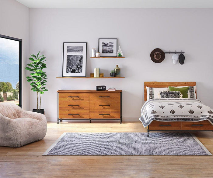 A modern bedroom featuring wooden furniture, a bed adorned with patterned bedding, a large window allowing natural light to pour in, and a soft chair for added comfort. A dresser provides storage, while wall art and floating shelves such as the Hasse 35″ Shelf enhance the decor. A cozy rug and a potted plant bring warmth and inviting style to the space.