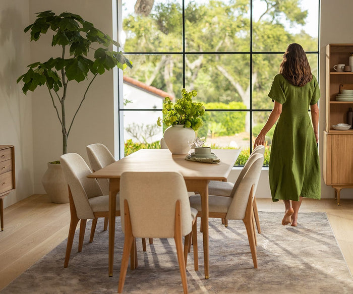 A woman in a green dress stands barefoot by large windows in a modern dining room with a wooden table, potted plants, and cream Hendrick Dining Chairs in White Oak featuring comfortable cushioned seats. Sunlight fills the room.
