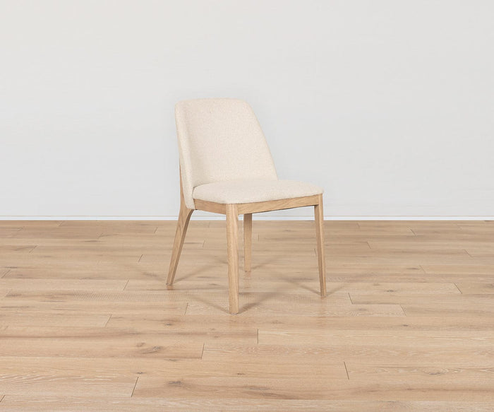 A Hendrick Dining Chair - White Oak, featuring a white oak frame and a cushioned seat upholstered in light beige, sits on a light wood floor against a plain white wall.