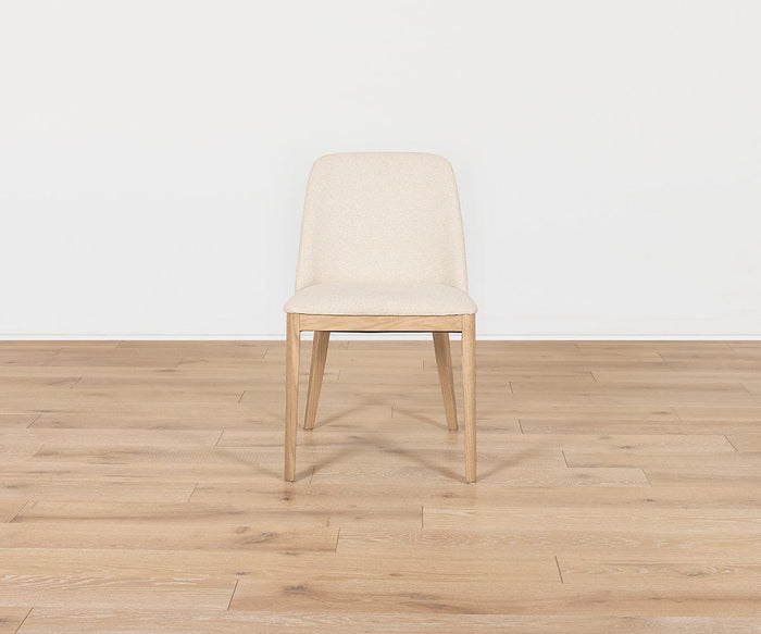 Centered on a light wood floor against a plain white wall, the Hendrick Dining Chair – White Oak features simple, light beige upholstery with a cushioned seat and a sturdy white oak frame.