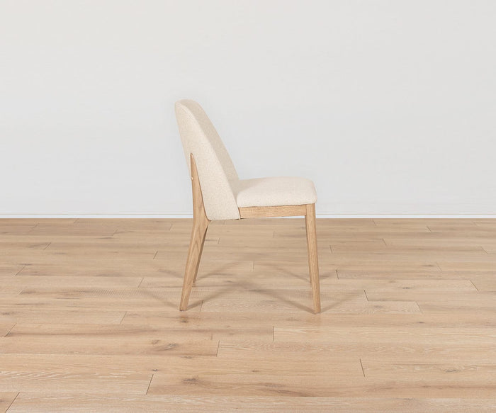 The Hendrick Dining Chair - White Oak is shown in side profile, highlighting its modern design with a white oak frame and light beige cushioned seat and backrest, placed on a light-colored wooden floor against a plain white wall.