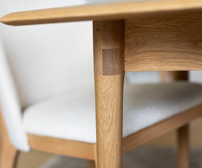 Close-up of a wooden table leg and edge, highlighting smooth craftsmanship and precise joinery. In the background, a blurred Hendrick Dining Chair - White Oak with a cushioned seat is partially visible.