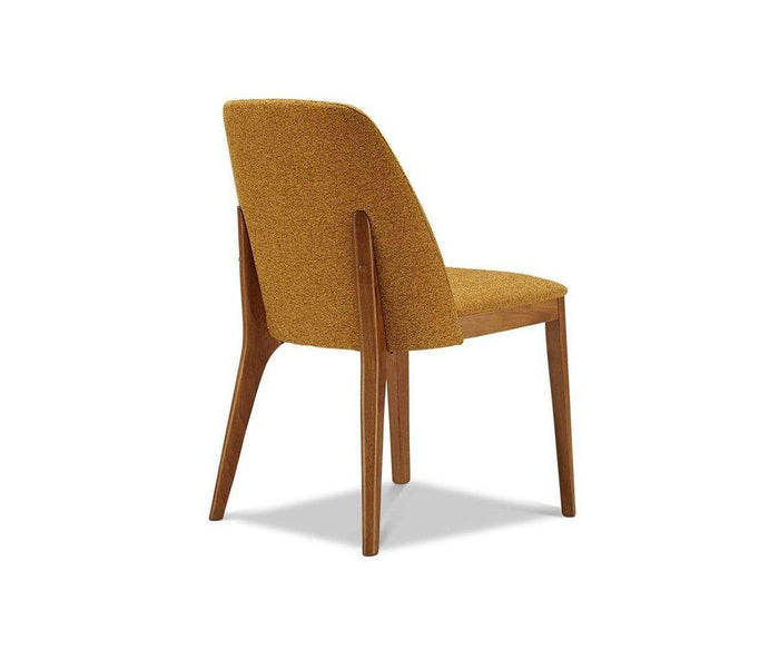 The Hendrick Dining Chair, seen from behind at an angle, showcases its modern wooden design with a curved backrest, four sturdy legs, and mustard yellow upholstery. With a durable frame and cushioned seat for comfort, this chair stands out against a plain white background.
